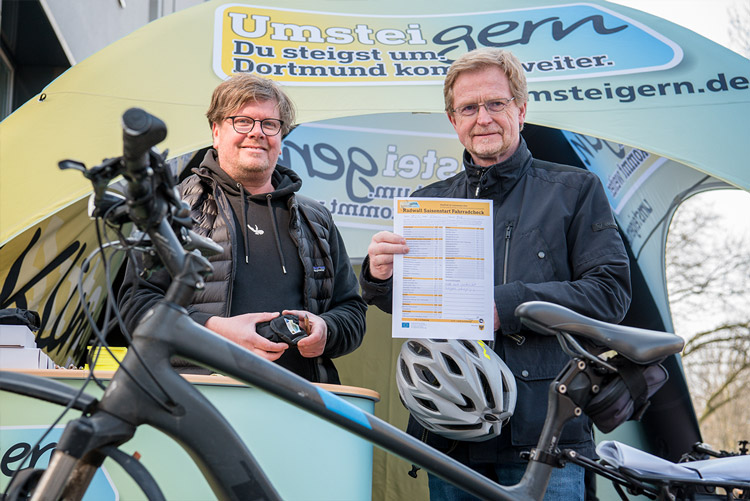 Zwei Männer stehen hinter einem Fahrrad und halten lächelnd eine Checkliste in die Kamera.