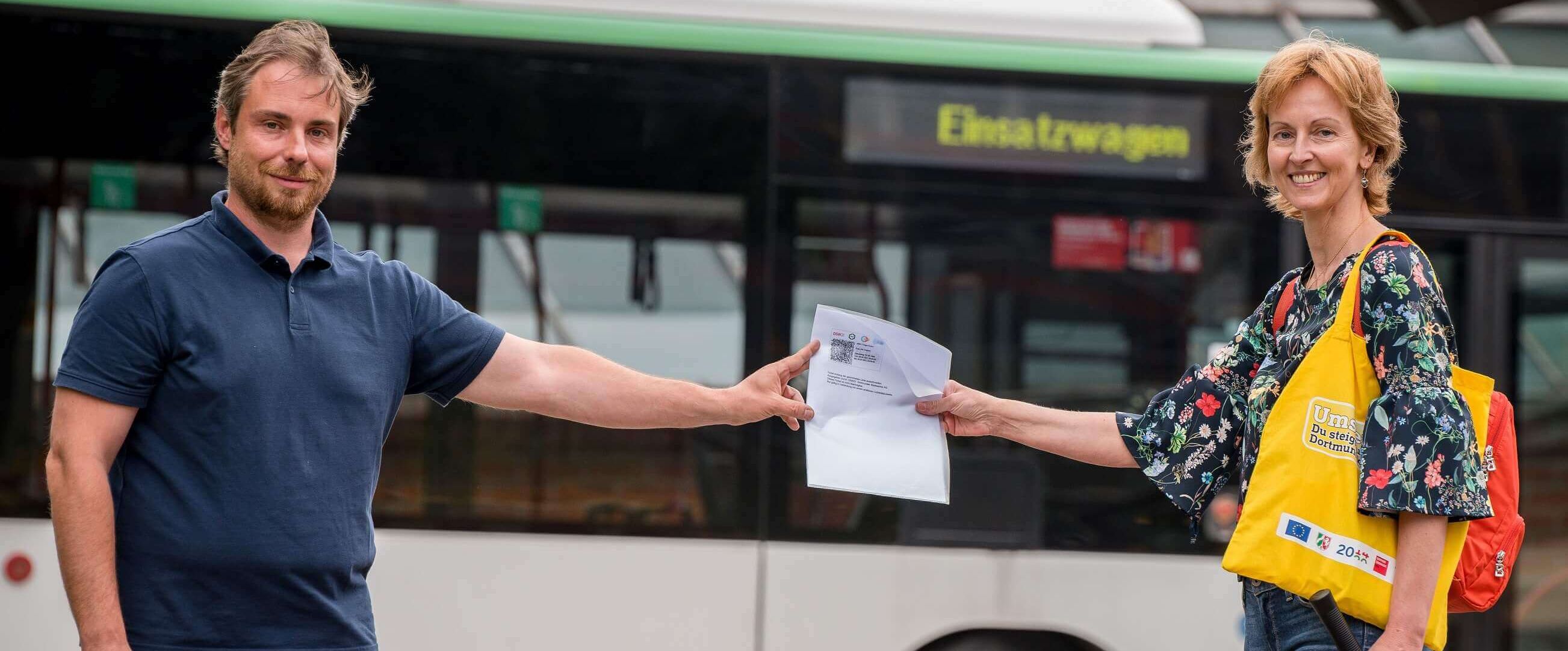 Eine Frau, welche einem Mann, ein Ticket anreicht. Im Hintergrund ist ein Linienbus abgebildet.